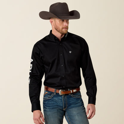 Man wearing a black Ariat shirt with branding, jeans, and a cowboy hat on a beige background