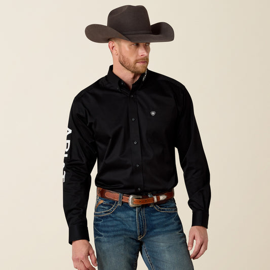 Man wearing a black Ariat shirt with branding, jeans, and a cowboy hat on a beige background