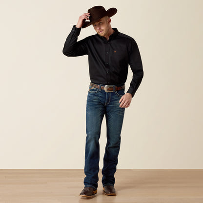 Man wearing a black Ariat shirt, blue jeans, and a brown cowboy hat on a beige background