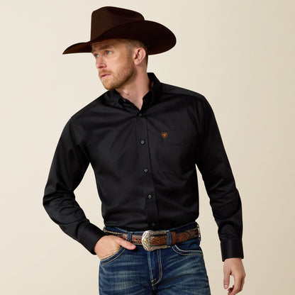 Man wearing a black Ariat shirt with a logo, blue jeans, and a brown cowboy hat on a beige background