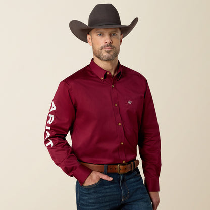 A man wearing a dark brown cowboy hat, a burgundy long-sleeve button-down shirt with white 'ARIAT' text on the sleeve, and blue jeans, standing against a solid beige background.