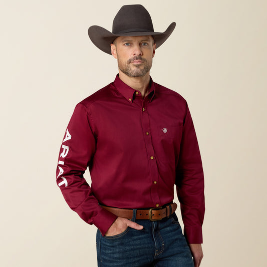 A man wearing a dark brown cowboy hat, a burgundy long-sleeve button-down shirt with white 'ARIAT' text on the sleeve, and blue jeans, standing against a solid beige background.