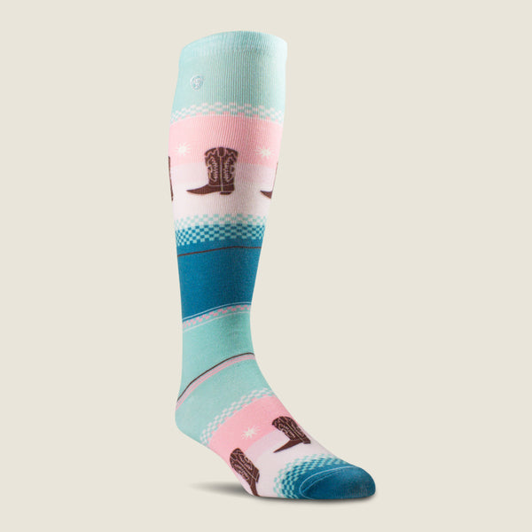 A single knee-high Ariat sock with a western boot theme, featuring a pattern of brown cowboy boots on pink and teal stripes over a white base, set against a solid light beige background.