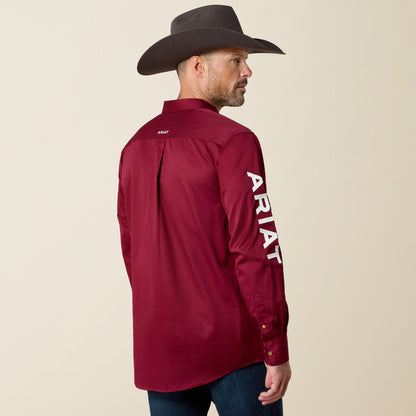 A man wearing a dark brown cowboy hat and a maroon long-sleeve western shirt with white vertical ARIAT branding on the sleeve, shown from the back against a light beige studio background.