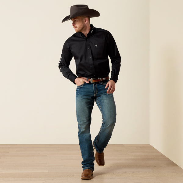 A man wearing a blackAriat  long-sleeve western-style button-down shirt, blue jeans, a brown cowboy hat, and brown boots, standing in a studio with a beige wall and light wood floor.