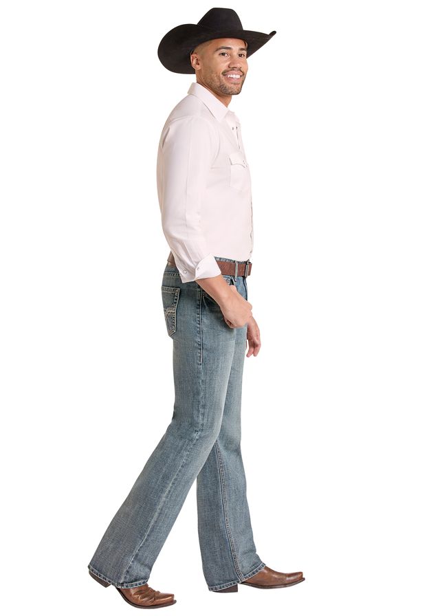 A man wearing a light pink western-style shirt, blue Rock & Roll Denim  jeans, a brown belt, and a black cowboy hat, against a plain white background.