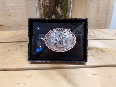 A belt buckle with pink bling and a barrel racer, oval in shape, with a rope edge, displayed on a black stand with a wooden background.