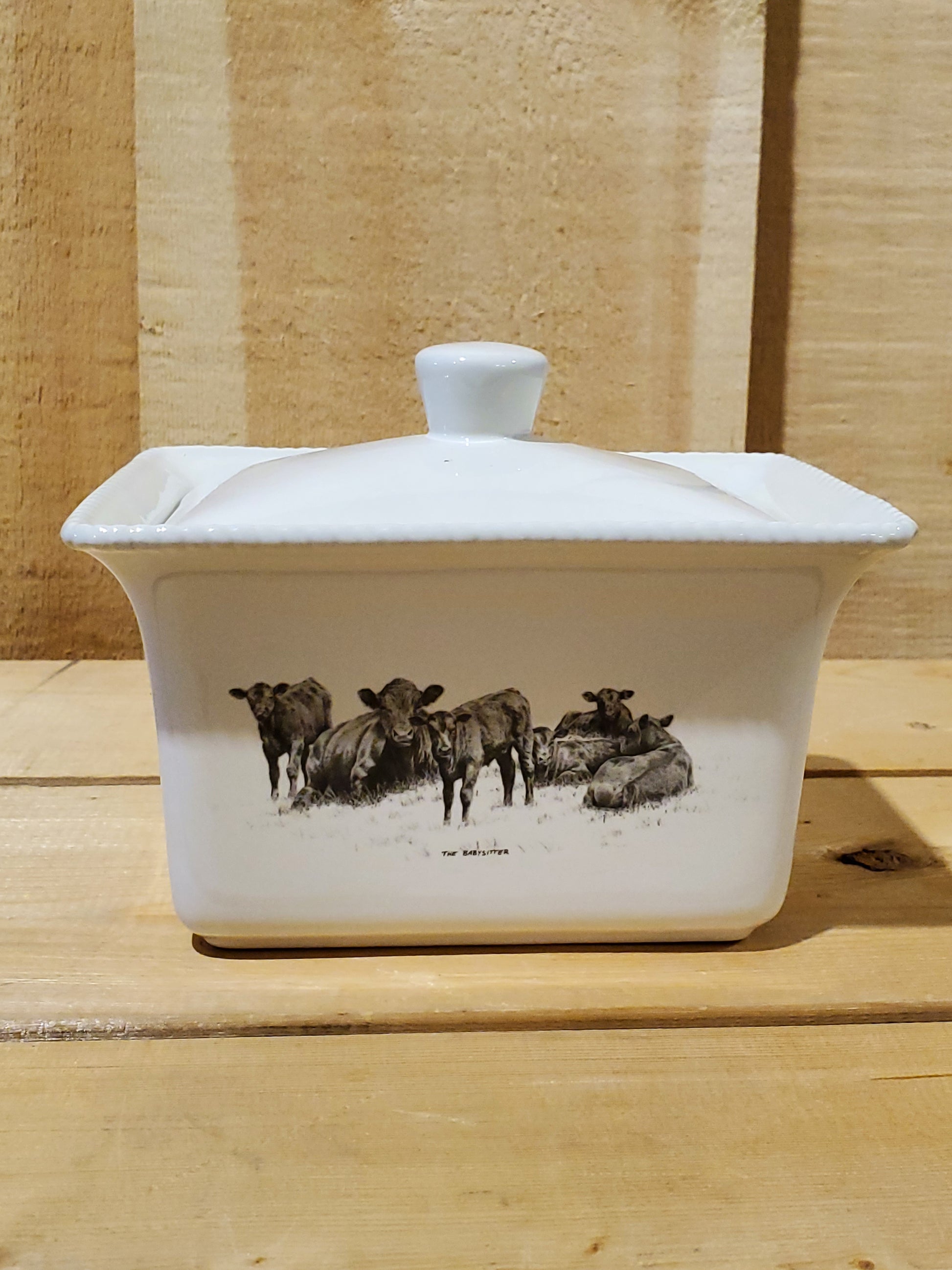 A white ceramic butter dish with a lid, featuring a black and white illustration of cattle on the side.