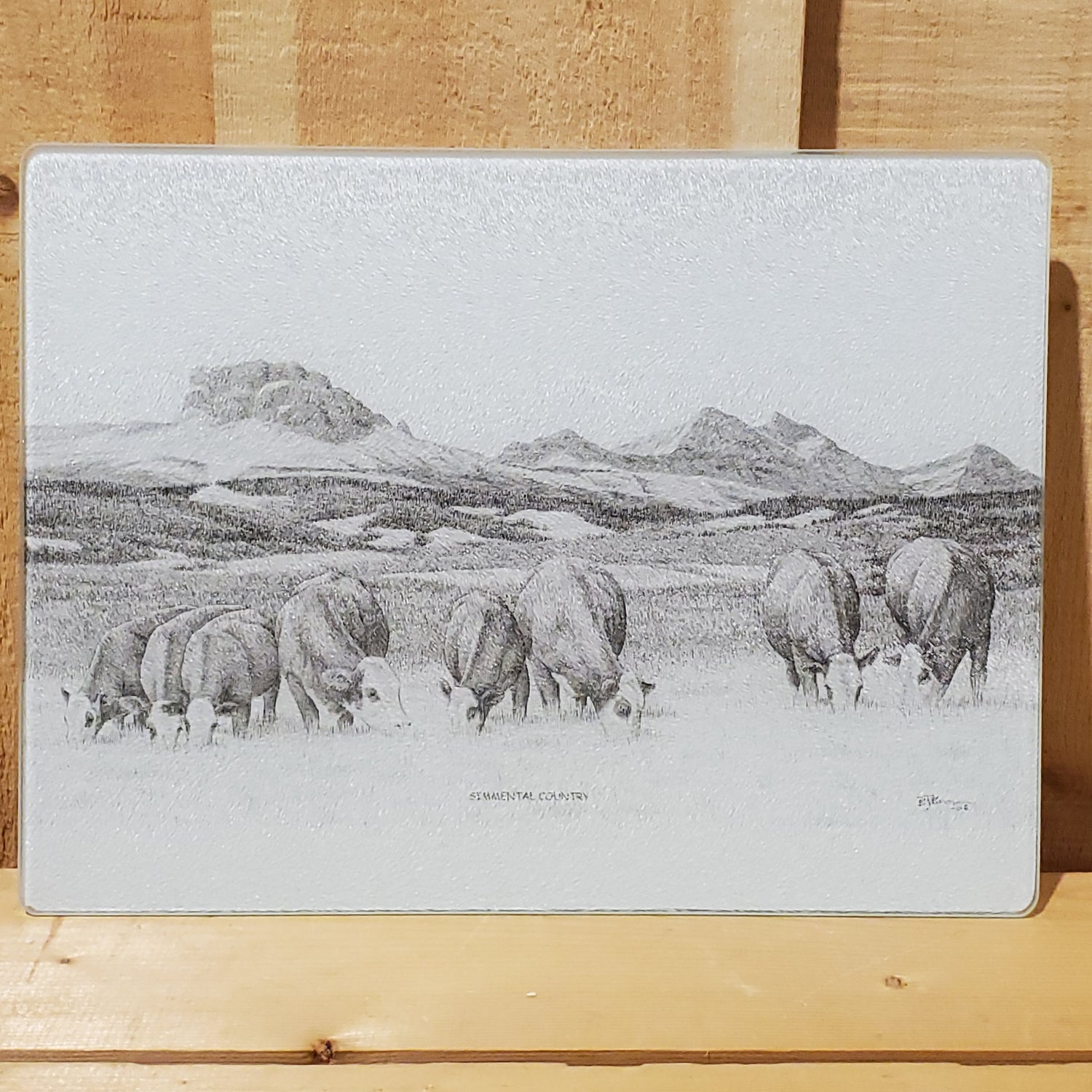 A glass cutting board with rubber feet featuring a black and white illustration of cattle in a field with mountains in the background.