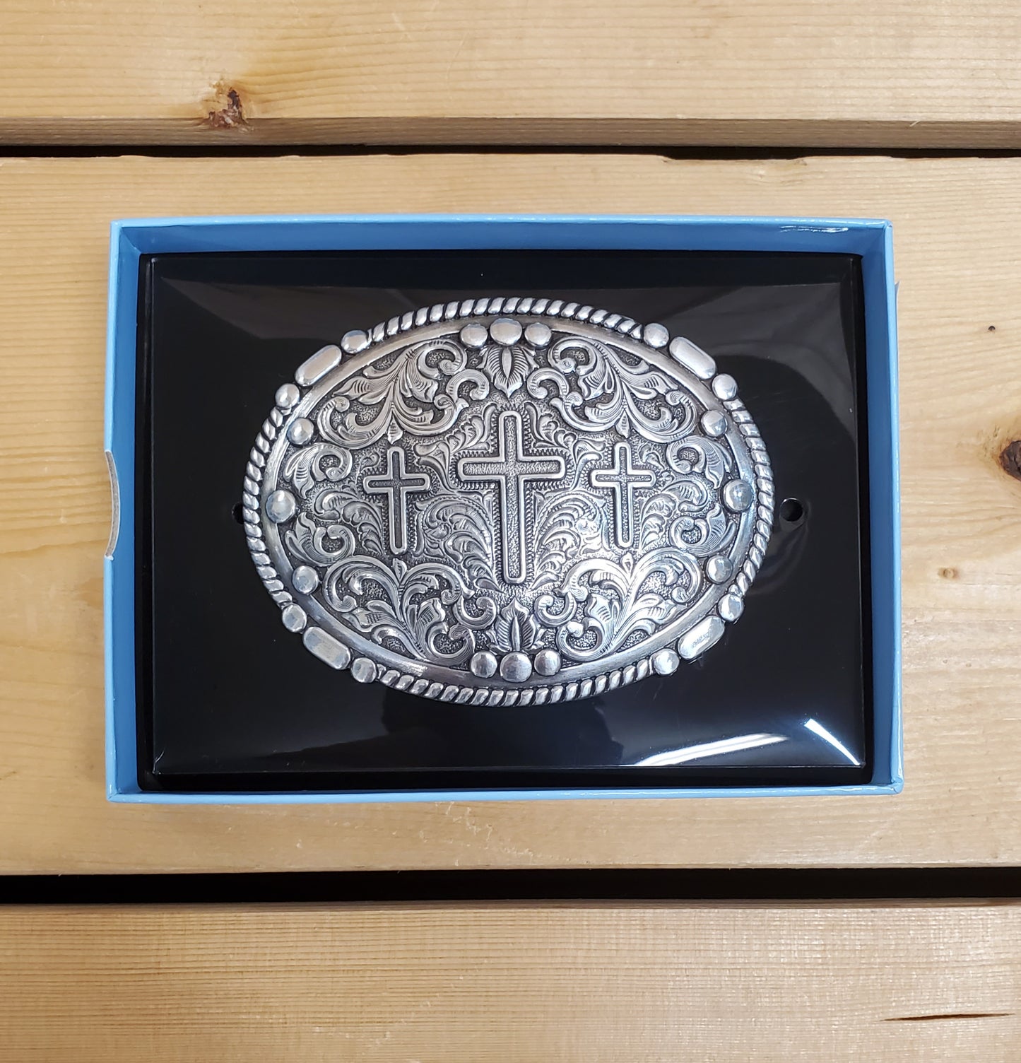 A metal belt buckle with an antique finish, featuring three cross figures and a scroll edge, displayed in a black and blue packaging.