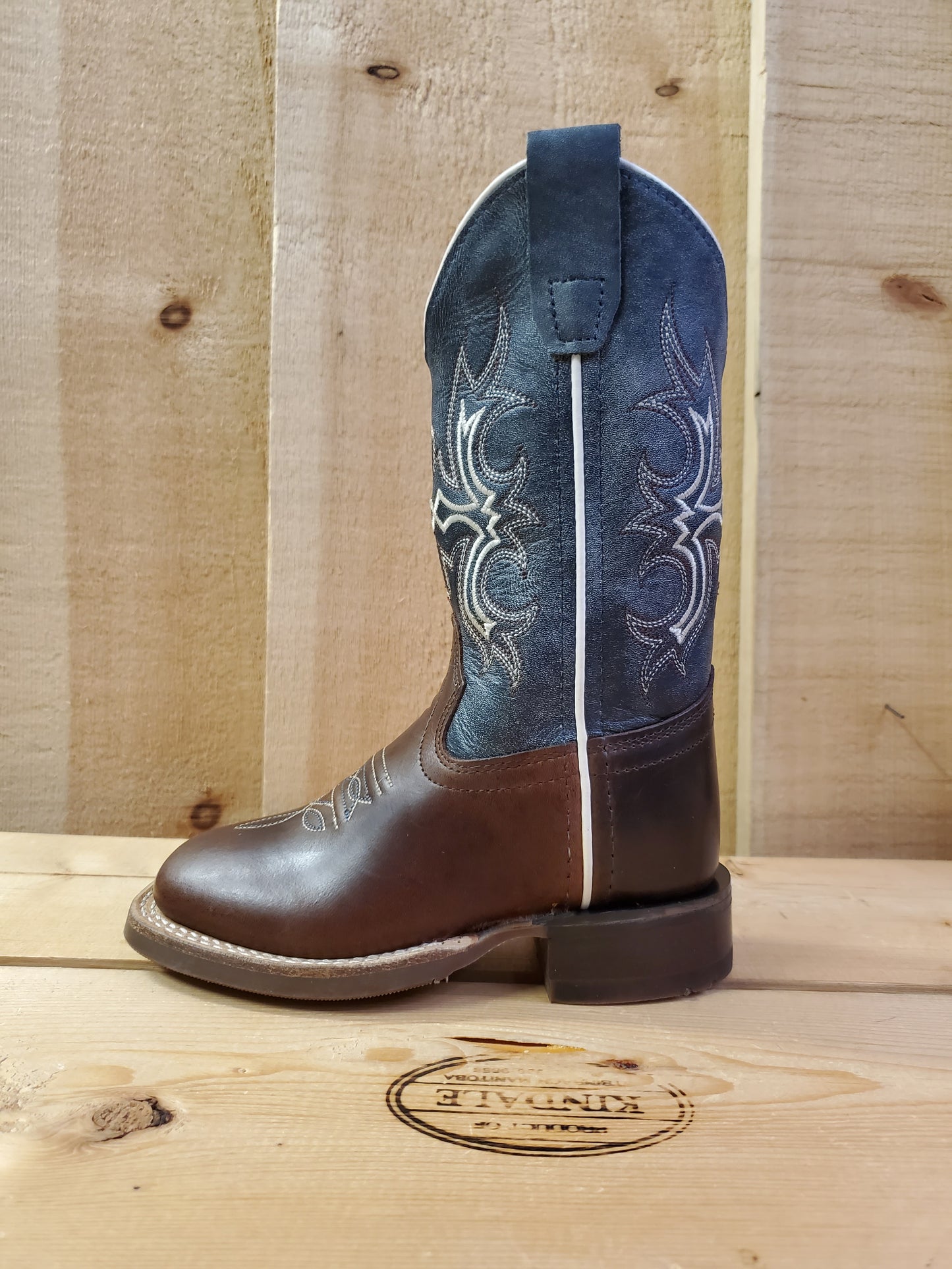 A kid's boot with a square toe, featuring white stitching on the shaft and a combination of navy blue and brown colors. 