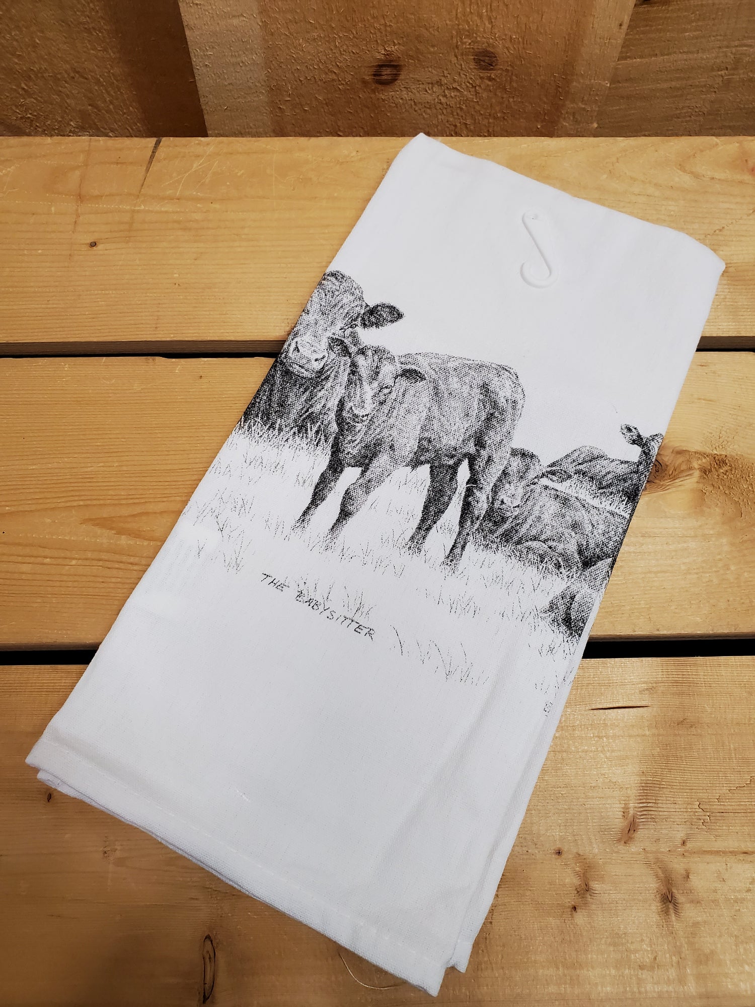 A white cotton tea towel with a printed black and gray cow design, laying on a wooden surface.