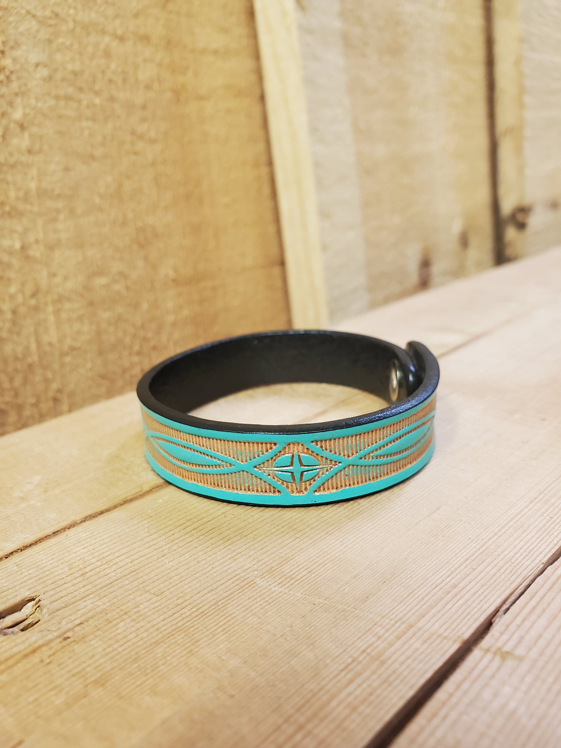 A teal leather bracelet with a stamped pattern, displayed on a wooden surface.