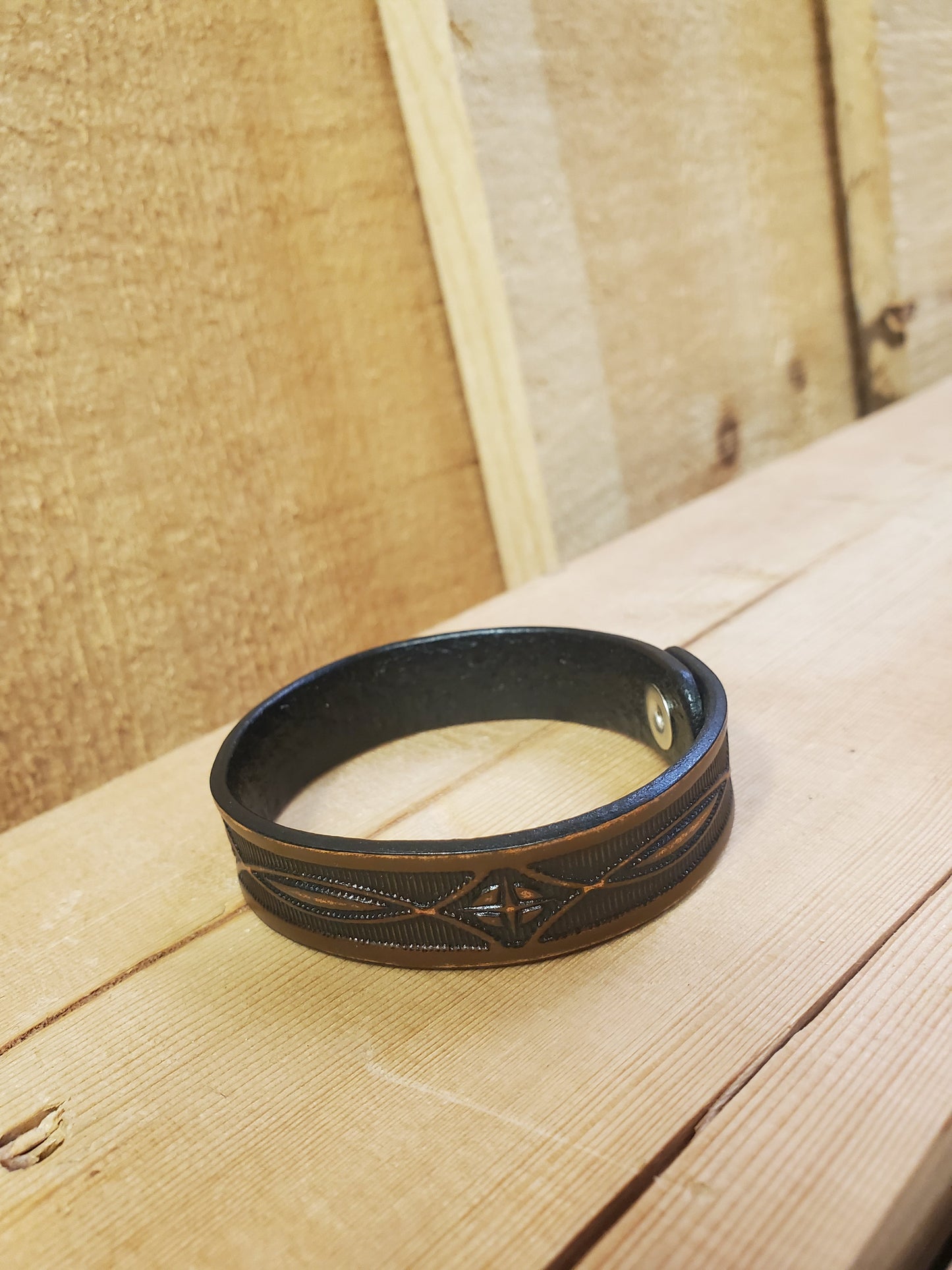 A brown leather bracelet with a stamped pattern, displayed on a wooden surface.