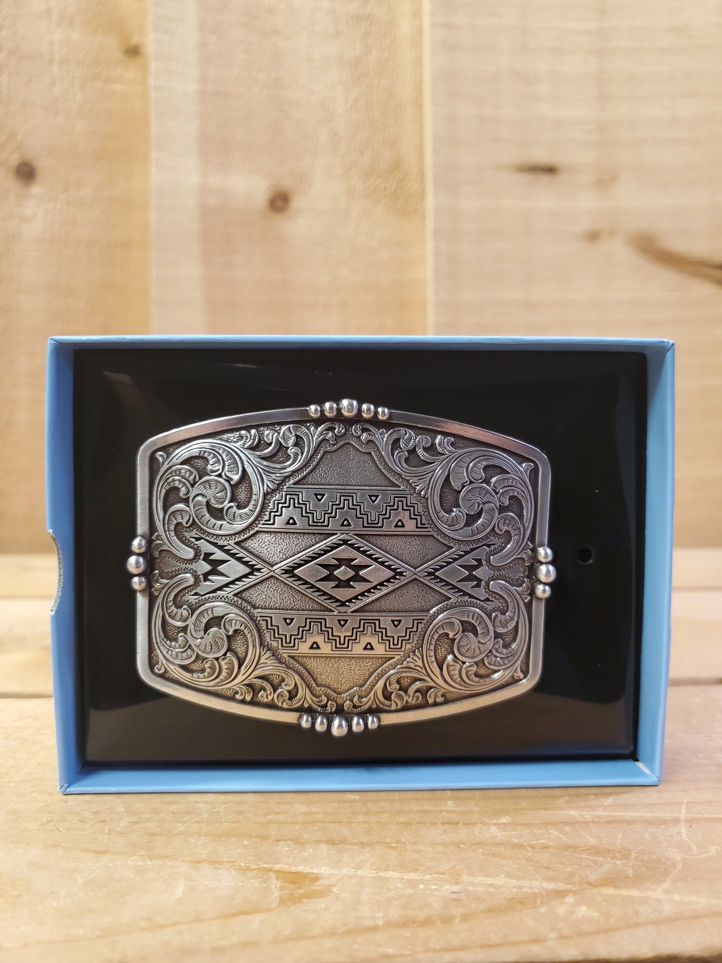 A belt buckle with a scroll and Southwest design, displayed in an open box with a black and blue interior on a wooden background