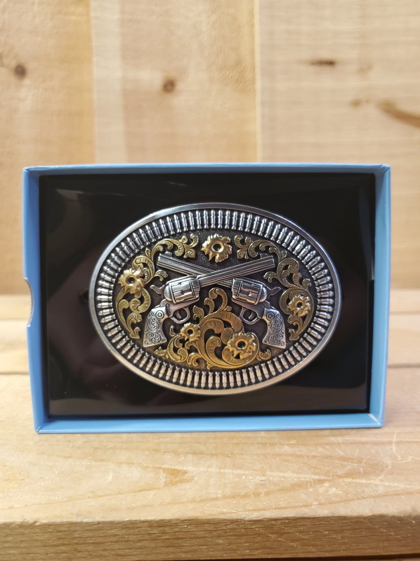 A 'Double Pistol' belt buckle by Nocona displayed in a box on a wooden background