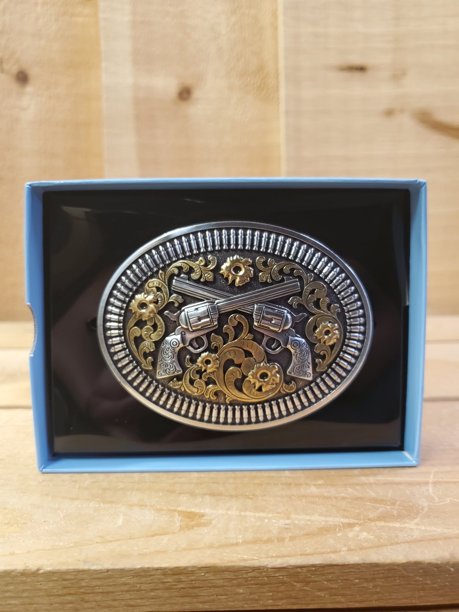 A 'Double Pistol' belt buckle by Nocona displayed in a box on a wooden background