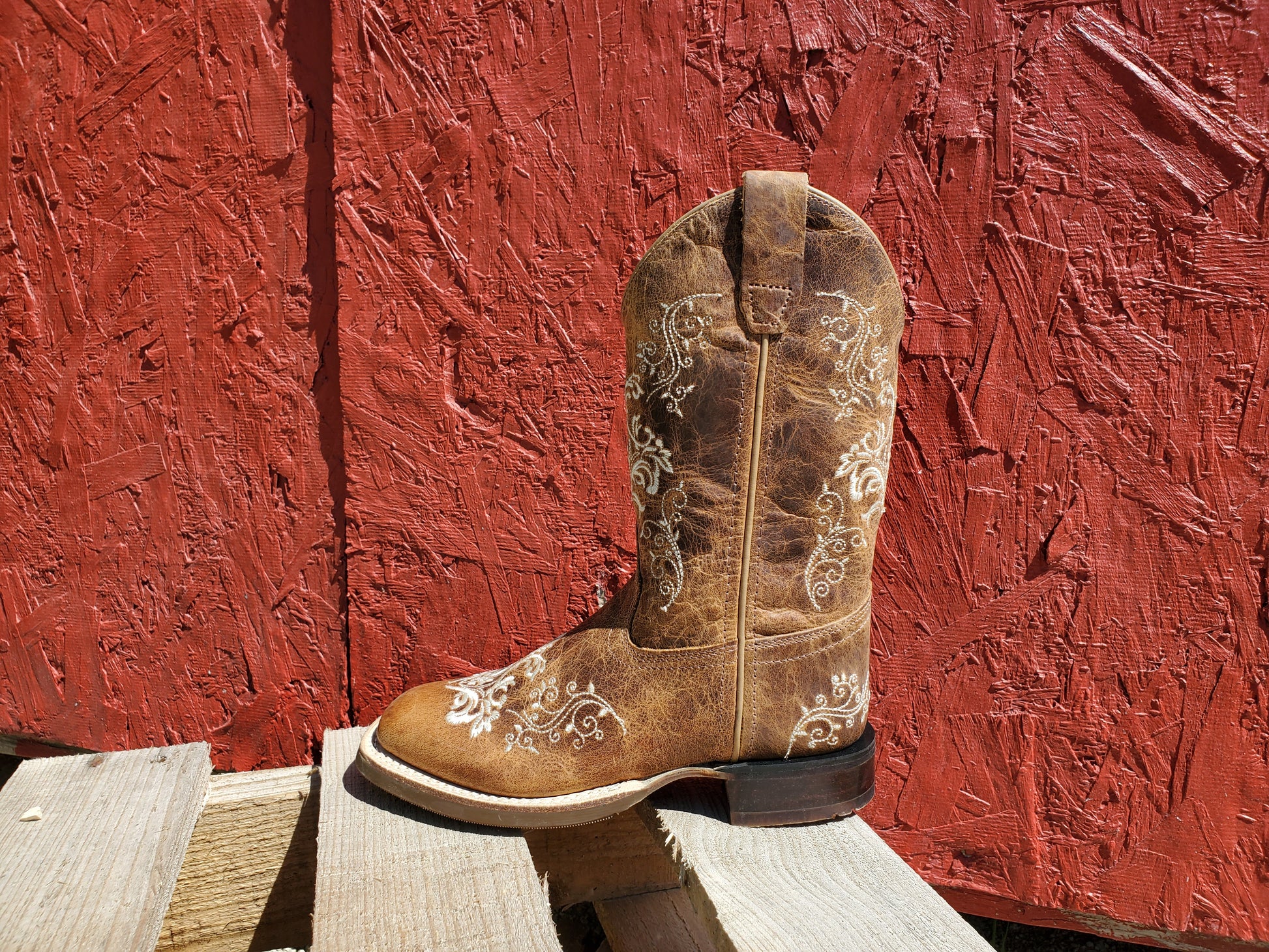 a brown kids cowboy boot with white floral stitching on a wooden surface with a red background
