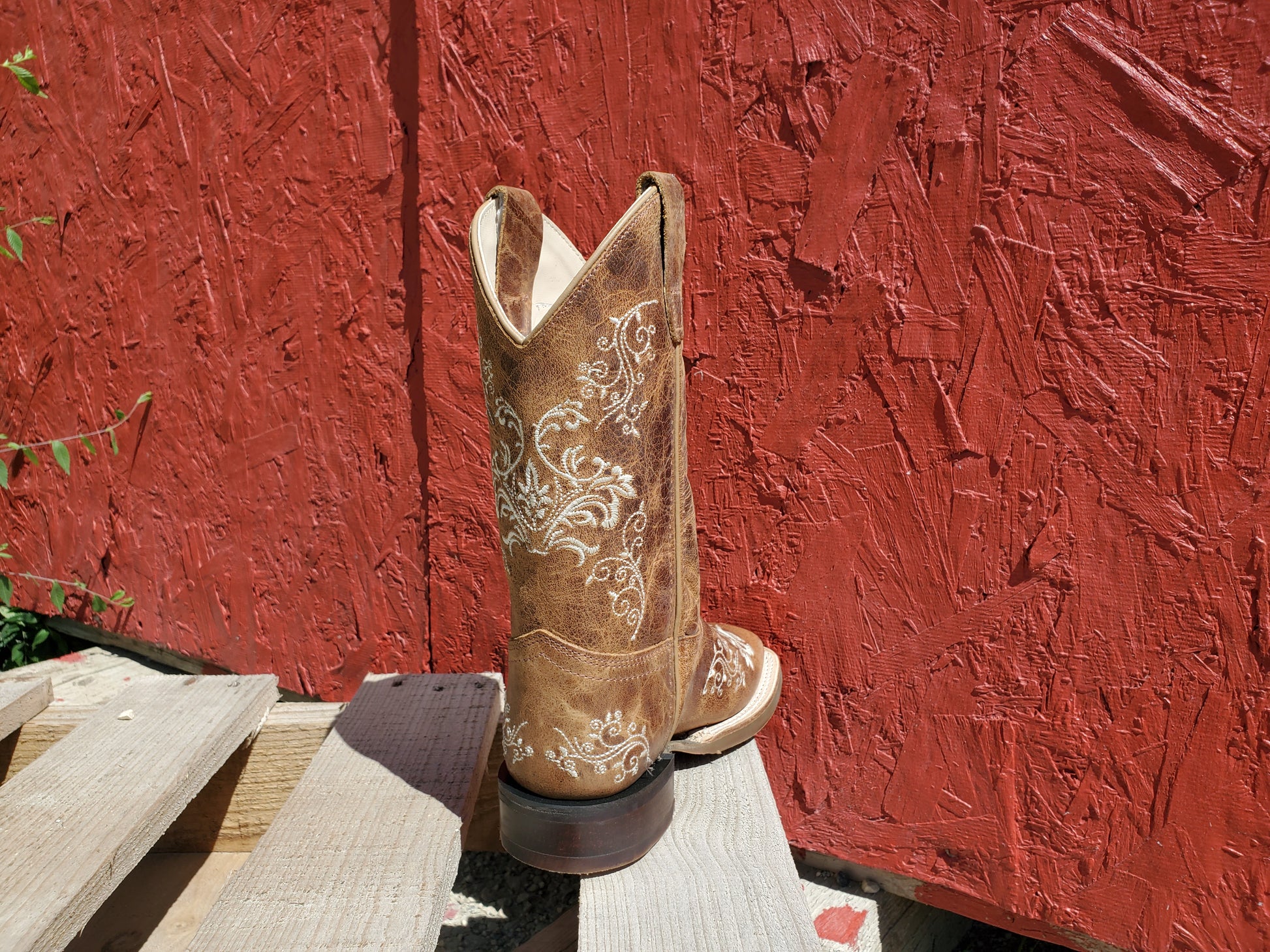 the back of a brown kids cowboy boot with white floral stitching on a wooden surface with a red background