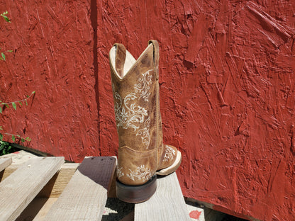 the back of a brown kids cowboy boot with white floral stitching on a wooden surface with a red background