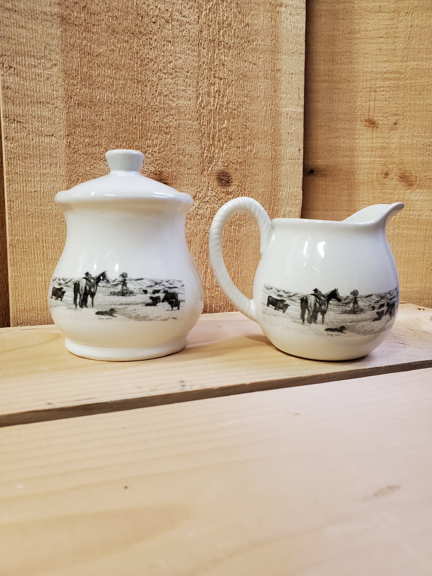 a white ceramic cream and sugar set with a black and white picture of a cowboy with his horse in a pasture with cows
