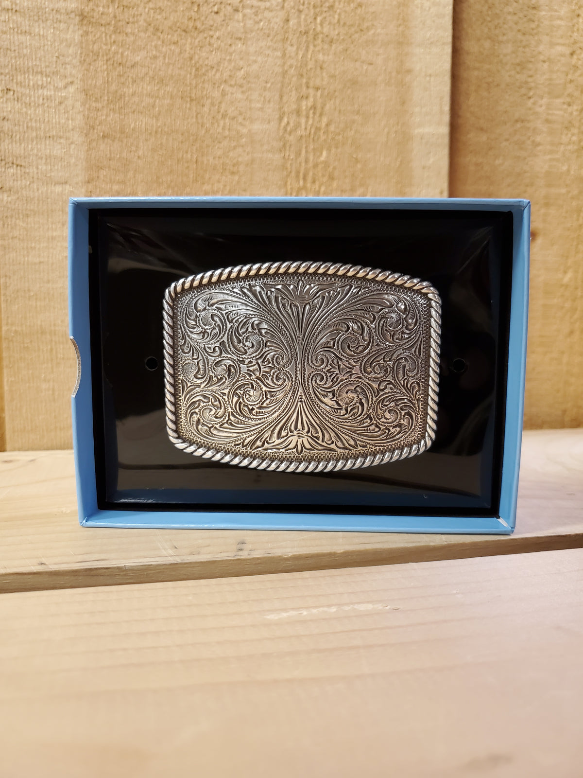 A belt buckle with scrollwork and rope edge detail, displayed in an open box with a blue interior.