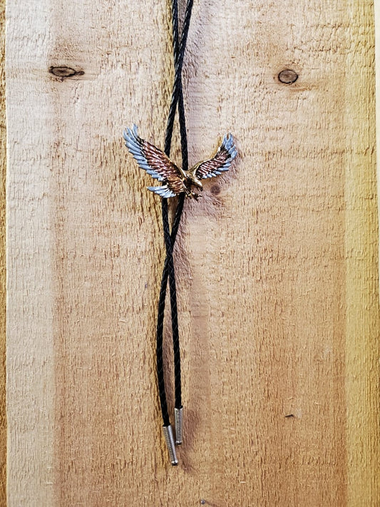 A bolo tie with a multicolored eagle pendant on a black cord, displayed against a wooden background.