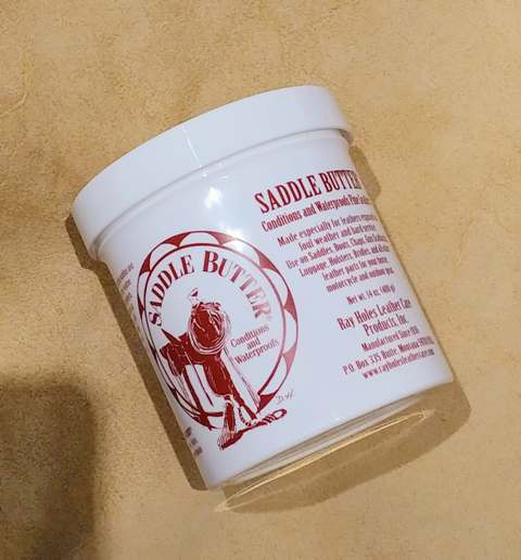 A container of Ray Hole's Saddle Butter, which is a solid white substance, a butter for conditioning and waterproofing leather products. on a tan background